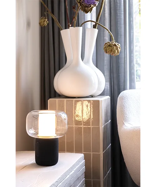 Лампа колонка_3 Компактна лампа с колонка Lux, поставена върху маса до книги и аксесоари.